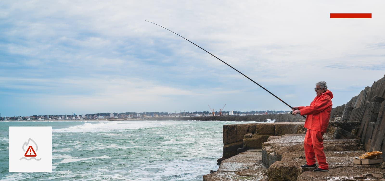 A rock fisherman up on the cliffs in an orange jacket and pants, leaning back with rod in hand, and tackle box by his feet